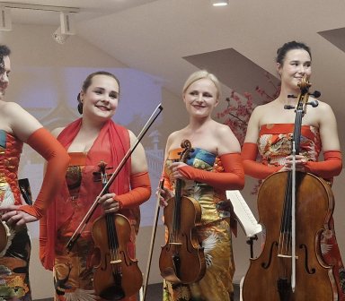 Kwartet smyczkowy. Kobiety trzymaja w rękach instrumenty i uśmiechają się do publiczności. Sa ubrane w kolorowe suknie.